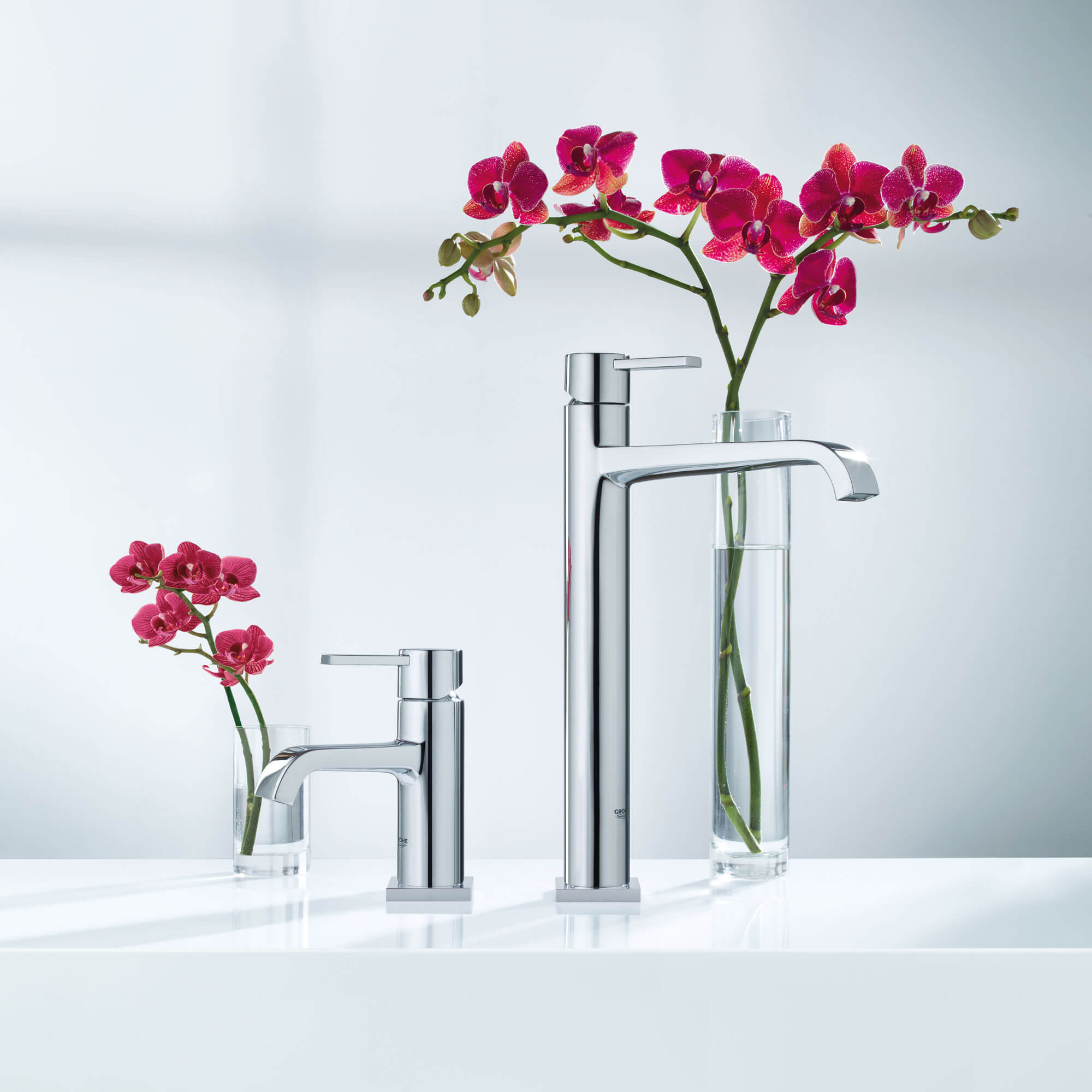 Two Grohe faucets next to two glasses filled with flowers.